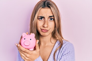 Beautiful hispanic woman holding piggy bank skeptic and nervous, frowning upset because of problem. negative person.