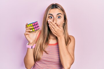 Beautiful hispanic woman holding colorful puzzle cube intelligence game covering mouth with hand,...
