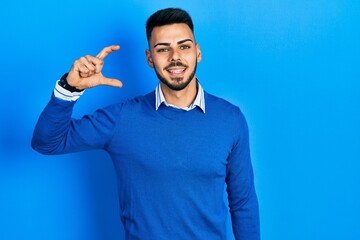 Naklejka premium Young hispanic man with beard wearing casual blue sweater smiling and confident gesturing with hand doing small size sign with fingers looking and the camera. measure concept.