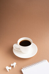 Vertical frame, a white cup with a saucer of strong black coffee, headphones and a notepad on a brown background