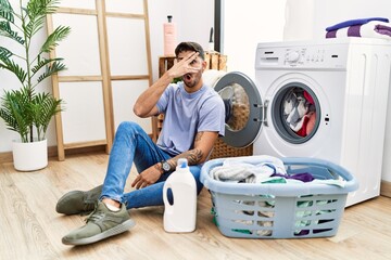 Young hispanic man putting dirty laundry into washing machine peeking in shock covering face and eyes with hand, looking through fingers with embarrassed expression.