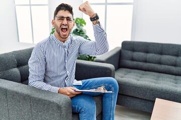 Young psychologist man at consultation office angry and mad raising fist frustrated and furious while shouting with anger. rage and aggressive concept.