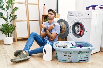 Young hispanic man putting dirty laundry into washing machine pointing to both sides with fingers, different direction disagree