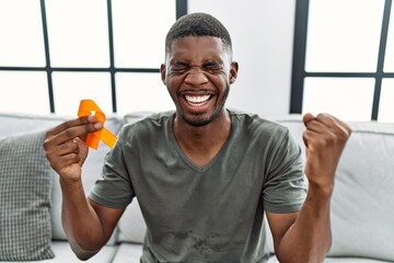 Young african american man holding awareness orange ribbon sitting on the sofa screaming proud,...