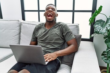 Young african american man using laptop at home sitting on the sofa angry and mad screaming...