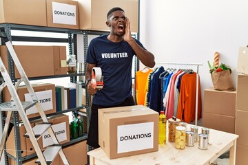Young african american volunteer man packing donations box for charity shouting and screaming loud to side with hand on mouth. communication concept.