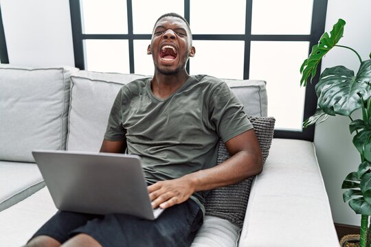 Young african american man using laptop at home sitting on the sofa angry and mad screaming frustrated and furious, shouting with anger. rage and aggressive concept.