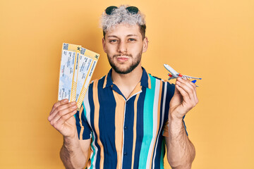 Young hispanic man with modern dyed hair holding plane toy and boarding pass puffing cheeks with...