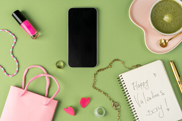 Cute pink cup with heart saucer and matcha latte on light green background and notepad, gift bag, accessories, girl's morning, top view. Mobile phone with black empty screen.