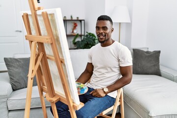 Young african man painting on canvas at home winking looking at the camera with sexy expression,...