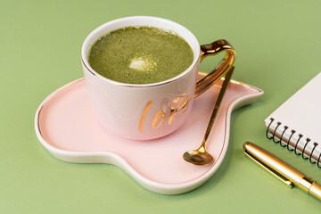 Cute pink cup with heart saucer and matcha latte and golden teaspoon on light green background and...