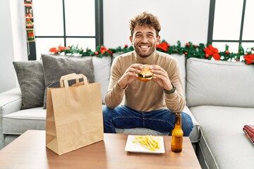 Young hispanic man eating take away hamburger sitting by christmas decor at home