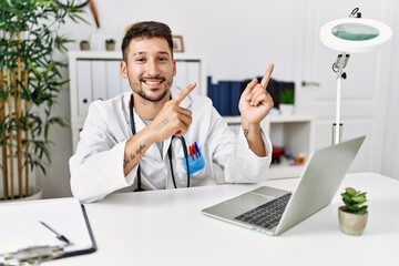 Young doctor working at the clinic using computer laptop smiling and looking at the camera pointing with two hands and fingers to the side.