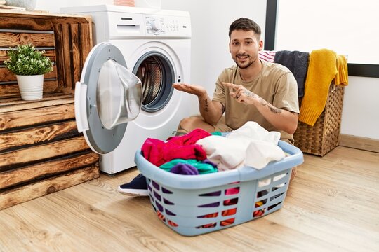 Young handsome man putting dirty laundry into washing machine amazed and smiling to the camera while presenting with hand and pointing with finger.