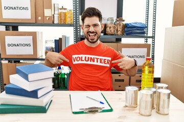Young man with beard wearing volunteer t shirt at donations stand winking looking at the camera...