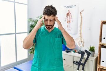 Young man with beard working at pain recovery clinic with hand on head for pain in head because stress. suffering migraine.