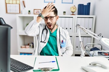 Young man with beard wearing doctor uniform and stethoscope at the clinic surprised with hand on...