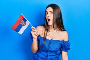 Young brunette teenager holding serbia flag scared and amazed with open mouth for surprise, disbelief face