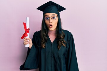 Young hispanic woman wearing graduation uniform holding diploma scared and amazed with open mouth...