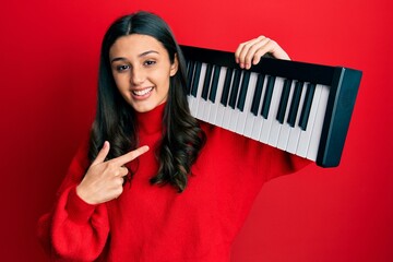 Young hispanic woman holding piano keyboard smiling happy pointing with hand and finger
