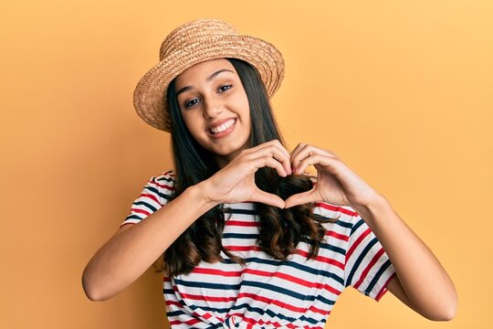 Young hispanic woman wearing summer hat smiling in love showing heart symbol and shape with hands. romantic concept.
