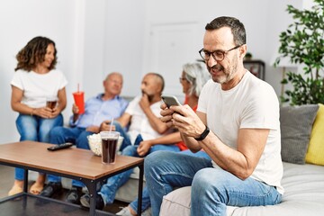 Group of middle age friends sitting on the sofa speaking. Man smiling happy using smartphone at home.