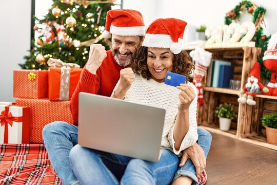 Middle Age Hispanic Couple Wearing Christmas Hat. Sitting On The Floor Using Laptop And Credit Card At Home.