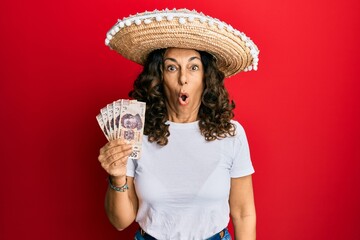 Middle age hispanic woman wearing mexican hat holding pesos banknotes scared and amazed with open mouth for surprise, disbelief face