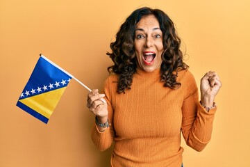 Middle age hispanic woman holding bosnia herzegovina flag screaming proud, celebrating victory and success very excited with raised arm - obrazy, fototapety, plakaty