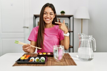 Obraz na płótnie Canvas Young brunette woman eating sushi using chopsticks smiling cheerful showing and pointing with fingers teeth and mouth. dental health concept.