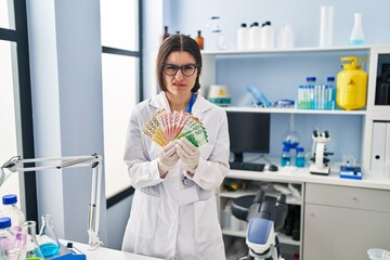 Young hispanic woman working at scientist laboratory holding money banknotes clueless and confused expression. doubt concept.