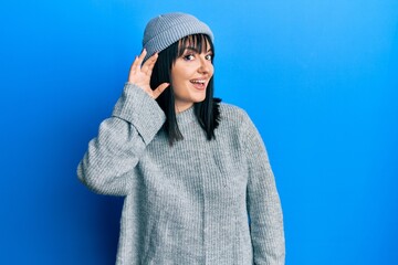 Young hispanic woman wearing wool sweater and winter hat smiling with hand over ear listening and hearing to rumor or gossip. deafness concept.