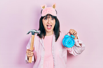 Young hispanic woman wearing sleep mask and robe holding alarm clock and hammer celebrating crazy and amazed for success with open eyes screaming excited.