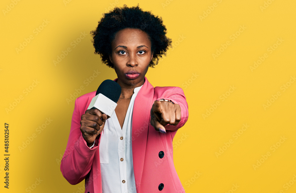 Wall mural African american woman with afro hair holding reporter microphone pointing with finger to the camera and to you, confident gesture looking serious