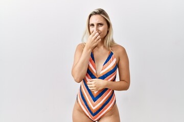 Young blonde woman wearing swimsuit over isolated background smelling something stinky and disgusting, intolerable smell, holding breath with fingers on nose. bad smell - obrazy, fototapety, plakaty