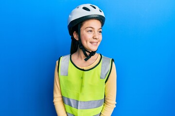 Beautiful brunette little girl wearing bike helmet and reflective vest looking away to side with smile on face, natural expression. laughing confident.