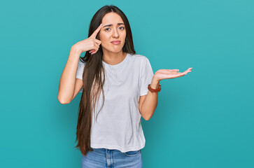 Young hispanic girl wearing casual white t shirt confused and annoyed with open palm showing copy space and pointing finger to forehead. think about it.