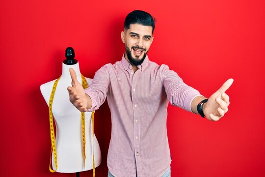 Handsome tailor man with beard standing by manikin looking at the camera smiling with open arms for hug. cheerful expression embracing happiness.