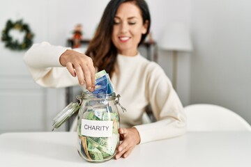 Middle age hispanic woman inserting banknote on charity jar sitting on the table by christmas decor at home
