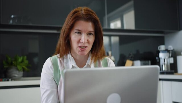 Cheerful woman chatting online via laptop, having zoom event with family or friends while sitting in kitchen, blurred background. Concept of technology