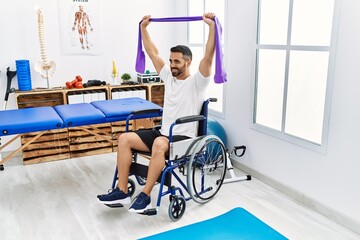 Young hispanic man patient having rehab session using elastic band sitting on wheelchair at clinic