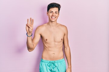 Fototapeta premium Young hispanic man wearing swimwear shirtless showing and pointing up with fingers number three while smiling confident and happy.