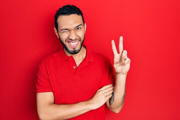 Naklejka premium Hispanic man with beard wearing casual red t shirt smiling with happy face winking at the camera doing victory sign. number two.