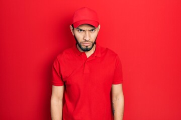Hispanic man with beard wearing delivery uniform and cap skeptic and nervous, frowning upset...