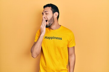 Hispanic man with beard wearing t shirt with happiness word message bored yawning tired covering mouth with hand. restless and sleepiness.