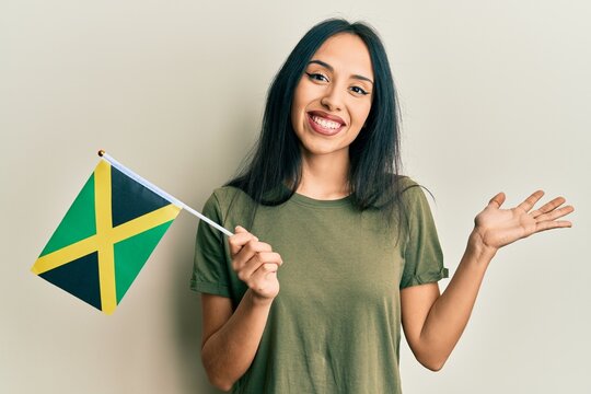 Young hispanic girl holding jamaica flag celebrating achievement with happy smile and winner expression with raised hand