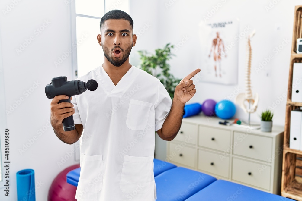 Wall mural Young indian physiotherapist holding therapy massage gun at wellness center surprised pointing with finger to the side, open mouth amazed expression.