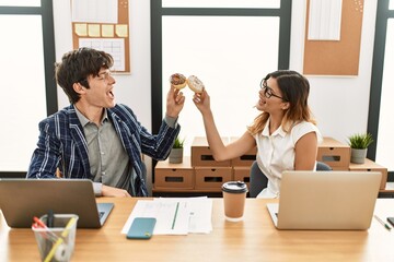 Two business workers smiling happy toasting with doughnuts at the office.