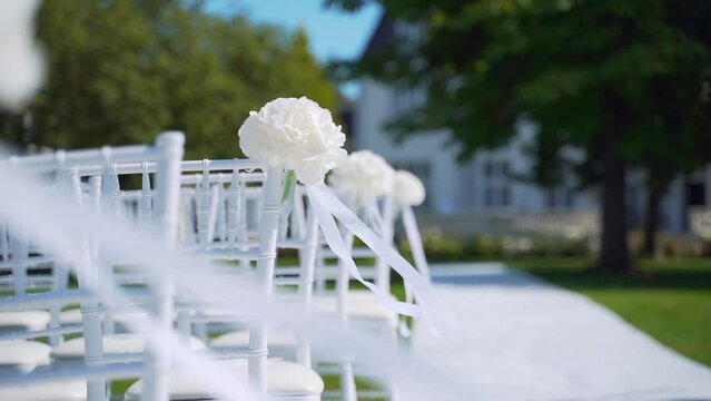 Close up. White chairs for guests at a luxury outdoor wedding ceremony. Event decoration with fresh flowers. Beautiful, convenient. armchair wind blows a ribbon on a lawn. Before celebration. Details