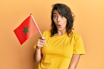 Young hispanic woman holding morocco flag scared and amazed with open mouth for surprise, disbelief face